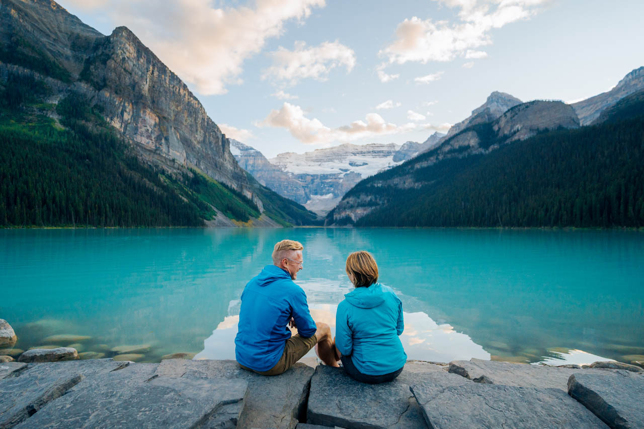 A couple sits on the shore of a turquoise lake staring out at a glacier in the distance while visiting Lake Louise at sunset