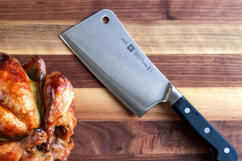 A large professional meat cleaver on a wooden cutting board with a rotisserie chicken