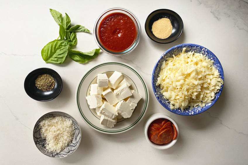 A flat lay of the ingredients needed to make a cheesy pizza dip: marinara sauce, cheese, fresh basil and pepperoni