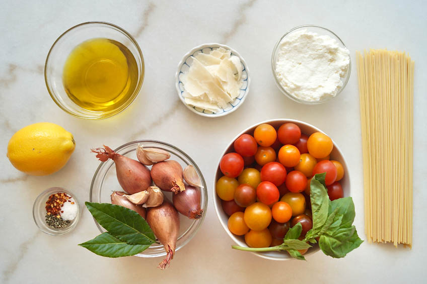 A flat lay of the ingredients needed for tomato confit pasta: cherry tomatoes, shallots, pasta, lemon, spices and cheese