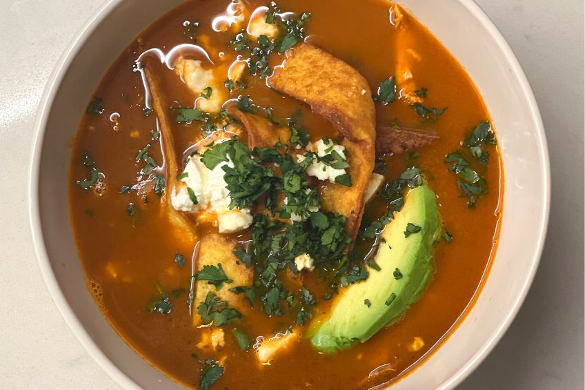 A bowl of Sopa Azteca with fried tortillas, fresh herbs, avocado and cheese