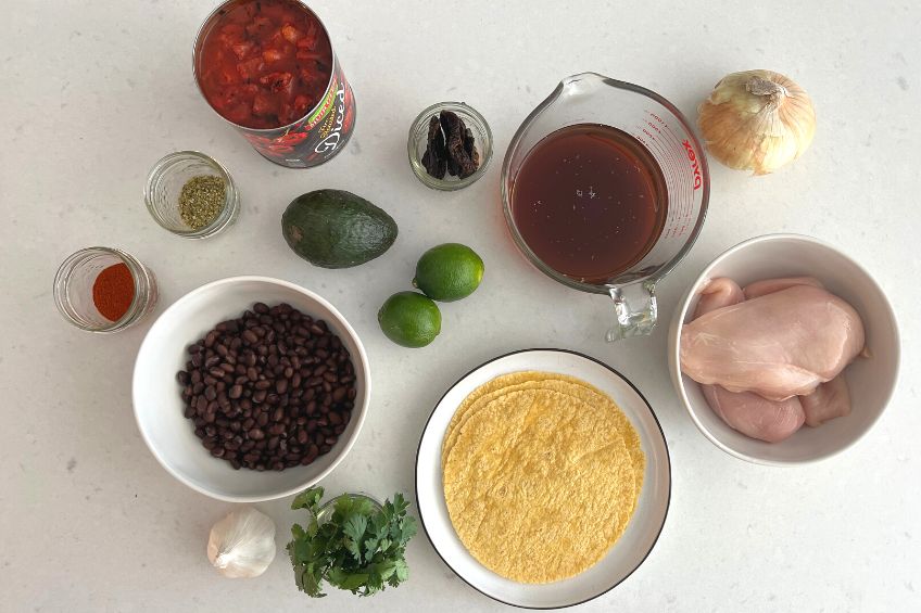 A flat lay of the ingredients needed for sopa azteca including tortillas, chicken breast, beans, canned tomatoes, and veggies