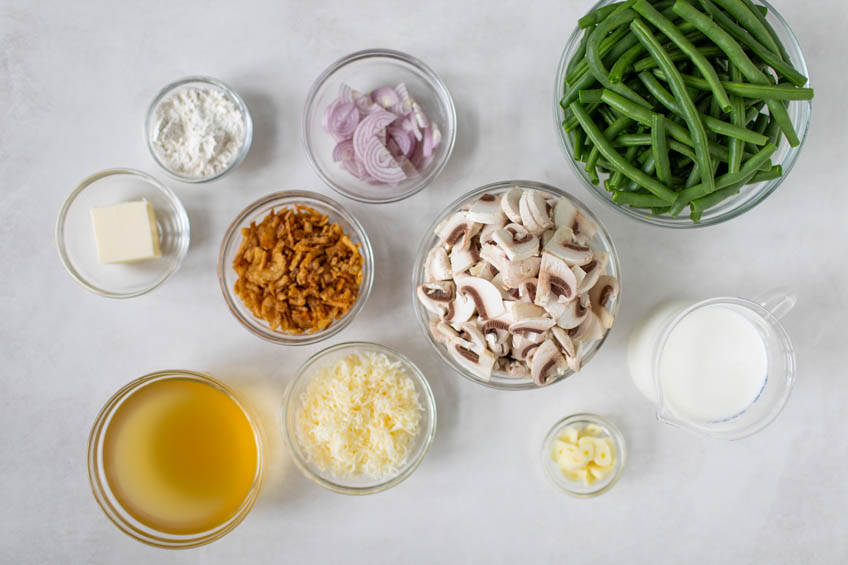 A flat lay of ingredients for a cheesyt green bean casserole: green beans, button mushrooms, stock, cheese, cream and more