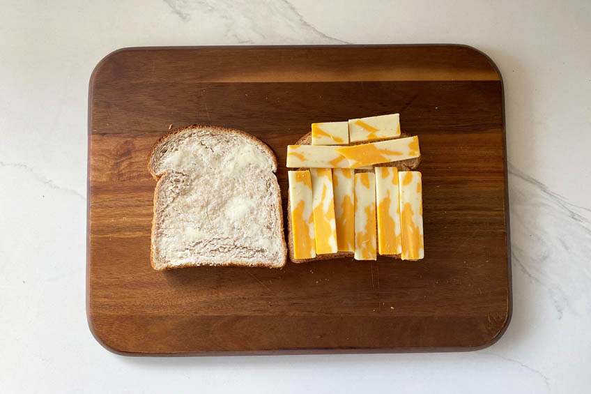 A wooden cutting board with a slice of buttered cheese and a second slice stacked with marble cheese slices
