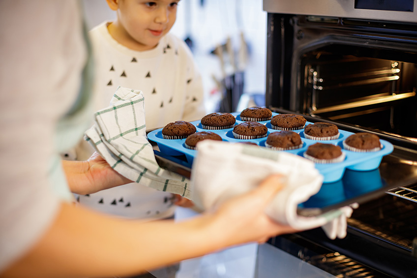 Mother taking baked muffins out of oven, her cute little boy is watching