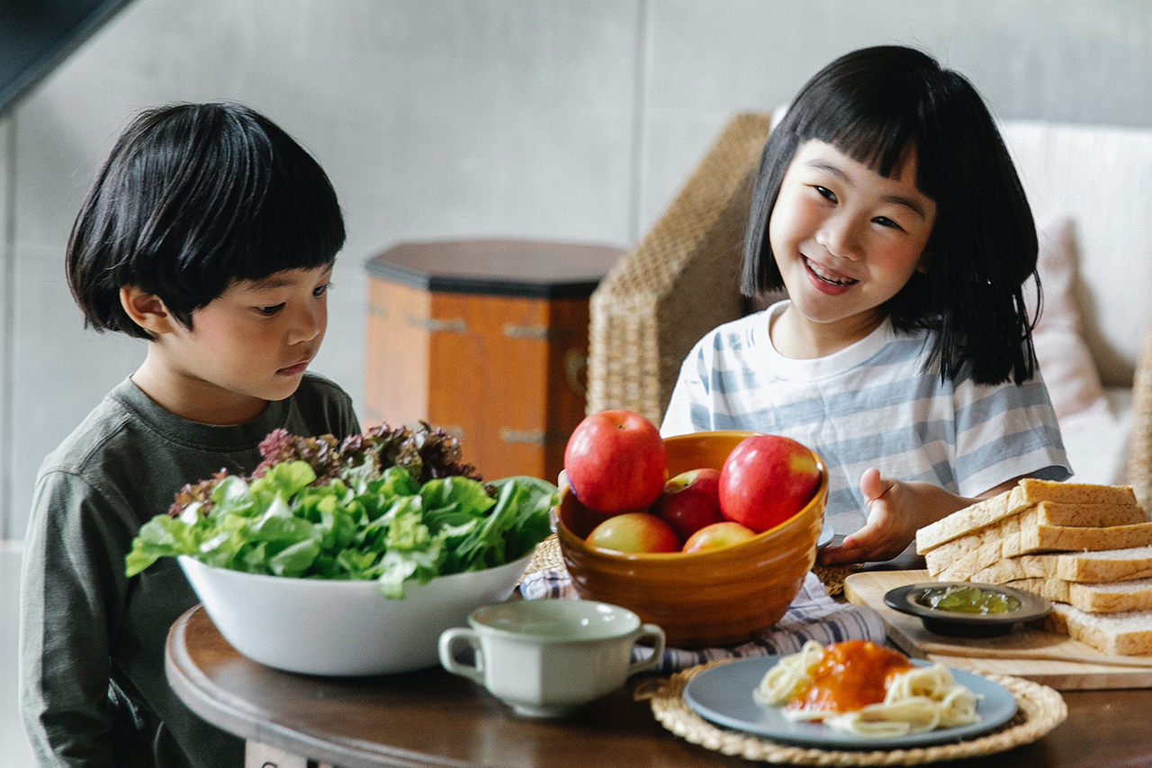 Two young kids sitting at a table with lots of vegetables, fruit and bread.