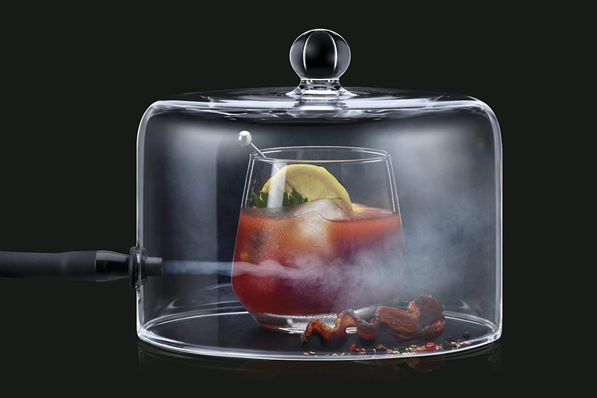 A caesar cocktail and a strip of bacon beneath a cloche, being infused with smoke.